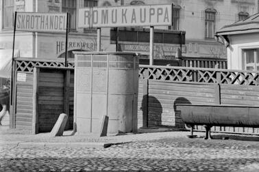 En oval pissoar av gjutjärn framför ett staket. Ovanför staketet finns skyltar med texten ROMUKAUPPA och SKROTHANDEL. I bakgrunden finns en stenbyggnad med en butik för herrkläder och en skoaffär. I förgrunden finns kullerstensbeläggning.