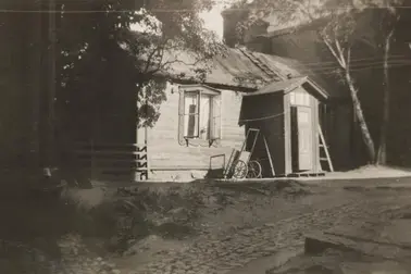 Yksikerroksinen pieni puurakennus pihapiirissä. Ikkunat ovat auki ja ovi raollaan. Talon seinustalla näkyvät maitokärryt. Piha on osittain kivetty.