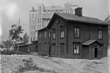Mitt i bilden står en träbyggnad i två våningar, och till vänster om den finns låga gårdsbyggnader. I bakgrunden reser sig ett stenhus i sex våningar som blivit klart samma år.