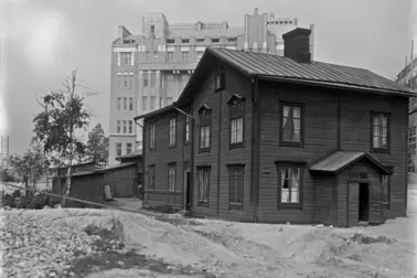 Mitt i bilden står en träbyggnad i två våningar, och till vänster om den finns låga gårdsbyggnader. I bakgrunden reser sig ett stenhus i sex våningar som blivit klart samma år.