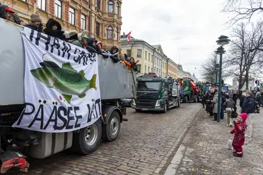 Abiturientit matkustavat penkkariasuihin pukeutuneina kuorma-autojen lavoilla. Ihmisiä on kerääntynyt seuraamaan penkkariajelua.