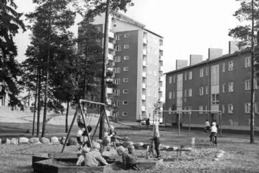 I förgrunden leker barn i en lekpark. En del av barnen är i sandlådan, några gungar och några har cyklar. I bakgrunden syns våningshus och träd. 
