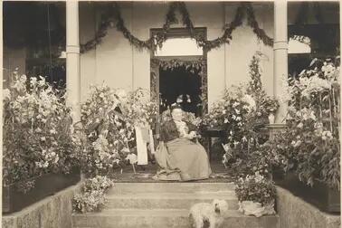 Aurora Karamzin fotograferad på sin 90-årsdag sittande framför ytterdörren till sin villa, omringad av blomplanteringar. En liten ljus hund sitter framför henne på trappan. 
