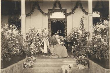 Aurora Karamzin fotograferad på sin 90-årsdag sittande framför ytterdörren till sin villa, omringad av blomplanteringar. En liten ljus hund sitter framför henne på trappan. 
