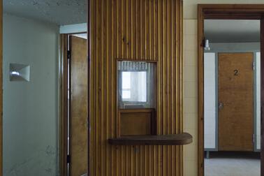 The toilet attendant’s workstation in a public toilet. A kiosk-like booth with a door and counter in front. The front wall is covered with vertical wood strips.