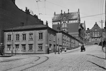 Nikolainkatu (= Snellmaninkatu ) 16, 18. Etualalla matala puutalo, sen takana uusia kivitaloja. Mukulakivikadulla ajaa mies hevosvaunuilla.