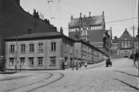 Nikolainkatu (= Snellmaninkatu ) 16, 18. Etualalla matala puutalo, sen takana uusia kivitaloja. Mukulakivikadulla ajaa mies hevosvaunuilla.