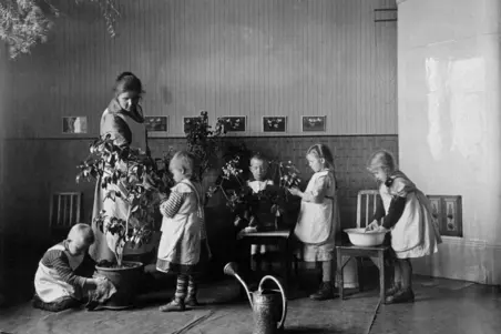 Fem barn sköter krukväxter under handledning av en kvinna i vitt förkläde. I högra kanten av bilden syns en vit kakelugn.
