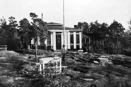 Ett envånings trähus omgiven av unga tallar med en flaggstång framför.