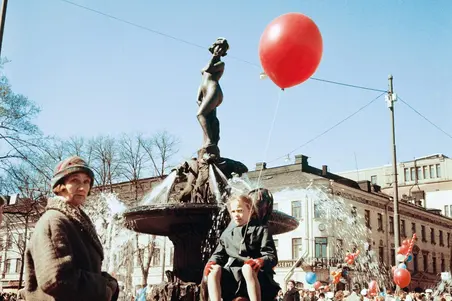 Havis Amanda på valborg. En flicka med en röd ballong i handen sitter på springbrunnens sälfigur. En kvinna står i förgrunden och tittar mot kameran. 