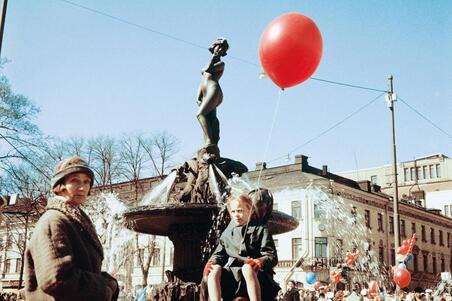 Suihkulähteen edustalla olevan hyljepatsaan päällä istuu tyttö, jolla on punainen ilmapallo. Vasemmalla etualalla seisoo nainen.