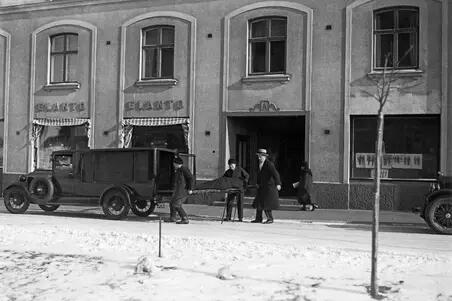 En snötäckt gata och trottoar framför ett höghus. Framför huset står en ambulans och annan bil. Tre män håller på att lyfta en en bår i ambulansens bakdel. I bakgrunden syns en kvinna som går förbi.