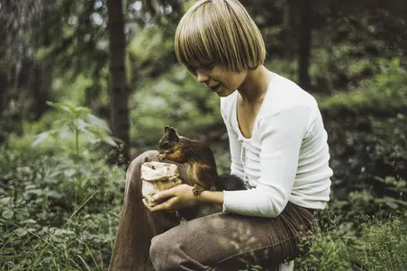 Hymyilevä poika on polvistunut maahan. Hänellä on valkoinen paita ja ruskeat vakosamettihousut. Hänen käsivarrellaan istuu orava, joka syö hänen kädessään olevasta paperipussista.