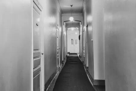 A black and white photo of an office corridor. Wooden doors on the left. Round lamps are hanging from the ceiling.