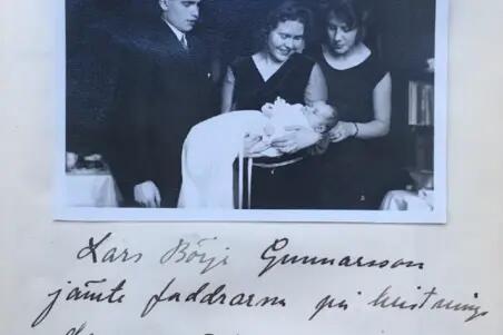 Kastemekkoon puettu lapsi naisen sylissä. Ympärillä seisoo mies ja nainen, kaikki katsovat lapseen.