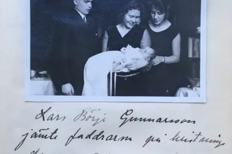 Kastemekkoon puettu lapsi naisen sylissä. Ympärillä seisoo mies ja nainen, kaikki katsovat lapseen.