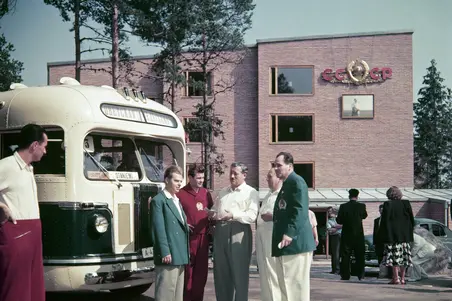 Neuvostoliittolaisia urheilijoita seisomassa bussin vieressä. Taustalla Otaniemen teekkarikylää, jonka seinälle on ripustettu Neuvostoliiton tunnus.
