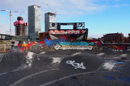 Suvilahti DIY-skeittipuisto. Puistossa betonisia ja asfalttipintaisia kumpareita ja ramppeja, jotka ovat kauttaaltaan värikkäiden graffitien peitossa. Kuvan taustalla kohoaa Kalasataman pilvenpiirtäjien rakennustyömaa.