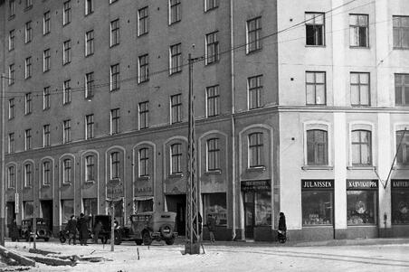 Seitsemänkerroksinen kivitalo kuvattuna kulman kohdalla kadulta. Talon edessä on autoja ja ihmisiä.