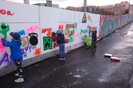 Nuoria lapsia maalaamassa Suvilahden graffitiseinällä. Lapset ovat piirtäneet nimiään ja yksinkertaisia hahmoja. Taustalla seinällä graffitiharrastajien monimutkaisempia teoksia.