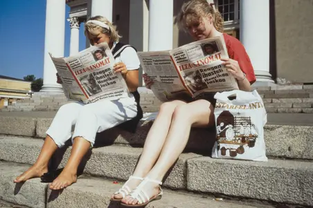 Två kvinnor sitter på domkyrkotrappan och läser Ilta-Sanomat. Intill den ena kvinnan står en plastkasse från Elanto. Den andra kvinnan är barfota. 