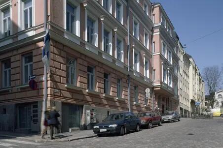 Vid hörnet av byggnaden går två mänskor. Tre bilar har parkerats längs med gatan. I bakgrunden syns andra 4-våningars byggnader. I ändan av gatan ett naket träd vars kvistar når upp till hustaket.