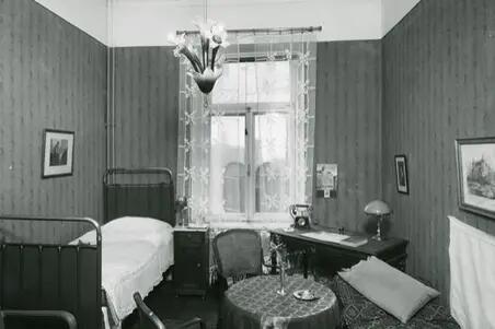 Till vänster syns en enkelsäng, till höger ett skrivbord med en skrivbordslampa och telefon. I mitten av rummet finns ett litet runt bord och två stolar.