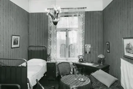Till vänster syns en enkelsäng, till höger ett skrivbord med en skrivbordslampa och telefon. I mitten av rummet finns ett litet runt bord och två stolar.