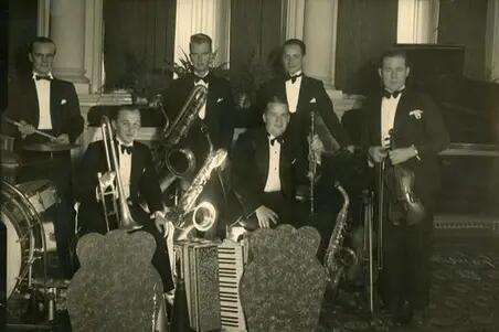 En smokingklädd sexmannaorkester med instrument: Trummor, trumpet, violin, saxofon och klarinett.