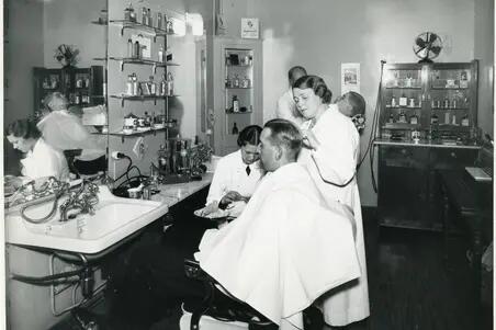 : I hotell Kämps barberarsalong. I förgrunden sitter en man i en frisörstol och blir klippt av en kvinna. En annan kvinna ger honom manikyr. I bakgrunden syns en annan kund och en manlig barberare.