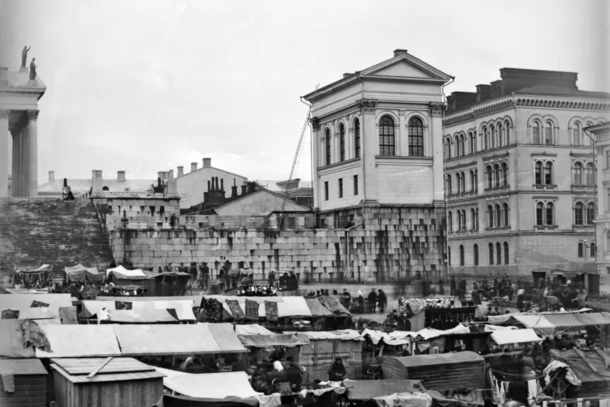 Senaatintori on täynnä markkinakojuja ja ihmisvilinää. Etualalla näkyy mm. rinkelikauppiaita myyntipöytiensä ääressä. Taustalla nousee Tuomiokirkon oikeanpuoleinen tasanne.