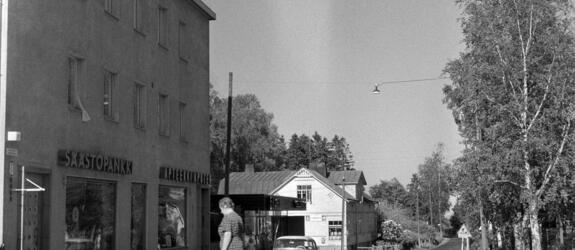 Tapanila, Veljestentie 17–21. Vasemmalla on kolmikerroksinen rapattu talo, jonka pohjakerroksessa on liikehuoneistoja. Seinässä on valomainokset SÄÄSTöPANKKI ja APTEEKKI APOTEK. Mekkoon pukeutunut nainen kävelee tien yli kohti rakennusta. Taustalla henkilöauto ja vaalea harjakattoinen puutalo, oikealla koivuja.