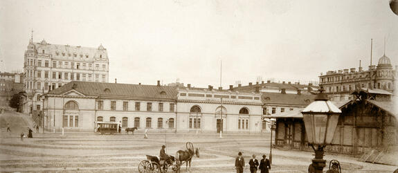 Hevosajureita rattaineen ja herrasväkeä Eteläsatamassa Pakkahuoneentorilla. Taustalla entinen sotaväen makasiini, vuodesta 1888 pakkahuone, Läntinen Rantakatu 10 (nyk. Eteläranta 10). Kadulla hevosvetoinen raitiotievaunu. Makasiinin takana kohoaa 1960-luvun lopulla purettu viisikerroksinen kivitalo osoitteessa Unioninkatu 14 - Eteläinen Makasiinikatu 3.