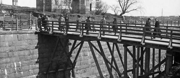 Människor promenerar på en träbro. Under bron löper hamnbanan. På andra sidan bron står en trävilla med en våning samt några kala björkar. I bildens vänstra kant syns Nationalmuseets torn.