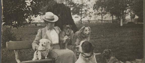 Kaksi naista puistossa. Toinen istuu penkillä, toinen seisoo hänen takanaan. Heidän kanssaan on viisi koiraa.