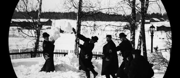 Män klädda i hattar och långa kappor leker med snöbollar. Bakom dem finns björkar och en gaslykta.