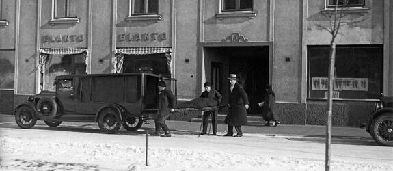 En snötäckt gata och trottoar framför ett höghus. Framför huset står en ambulans och annan bil. Tre män håller på att lyfta en en bår i ambulansens bakdel. I bakgrunden syns en kvinna som går förbi.