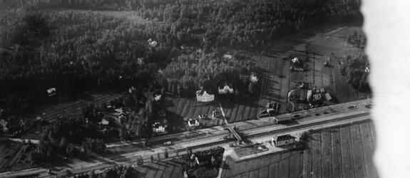 Keskellä kuvaa kulkee junarata, sen ympärillä on rakennuksia mutta enimmäkseen metsää ja peltoja. 