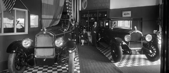 Autoliikkeessä on mustavalkoruudullinen lattia ja kaksi käytävämattoa. Tilassa on näytteillä avoauto ja umpiauto. Seinällä on Suomen lippu ja USA:n lippu. Kolme miestä seisoo takaseinän luona, ja heidän välissään on tuoli, jolla nainen istuu.