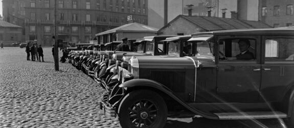 Vuokra-autoja siistissä rivissä mukulakivetyllä aukiolla. Taustalla vasemmalla kohoaa hotelli Seurahuoneen talo ja oikealla rakennuksia, jotka sijaitsivat ennen nykyisen Postitalon paikalla.