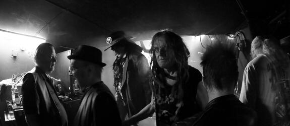 Rockmusiker i en dunkel bar. Mitt på bilden står Pelle Miljoona och tittar in i kameran. Bakom honom står Andy McCoy.
