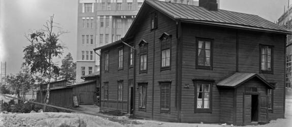 Kuvan keskellä on kaksikerroksinen puurakennus, jonka vasemmalla puolella on matalia piharakennuksia. Taustalla kohoaa samana vuonna valmistunut kuusikerroksinen kivitalo. 