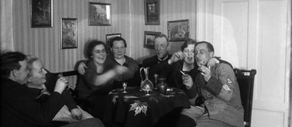 Seitsemän ihmistä istuu pyöreän pöydän ääressä. Joillakin on kädessä savuke ja snapsilasi. Pöydällä on snapsilaseja ja kaksi juomakaadinta. Yhdellä miehistä on suojeluskunnan univormu. 