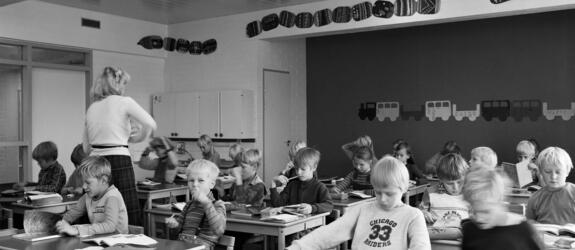 Alakoululaisia oppitunnilla luokassaan. Oppilaiden pöydillä on kirjapinoja, opettaja kiertää luokassa.