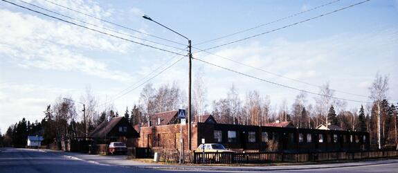 Tasakattoisia punatiilisiä rivitaloja, joiden takana näkyy harjakattoisia omakotitaloja. Etualalla tienristeys.