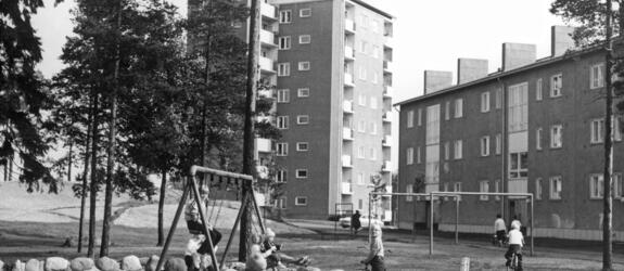 Etualalla leikkipuistossa leikkiviä lapsia. Osa lapsista on hiekkalaatikolla, muutama kiikkuu ja parilla on polkupyörät. Leikkipuisto on kivistä kasatun aidan sisälle jäävä hiekkakenttä. Taustalla näkyy kerrostaloja ja puita.