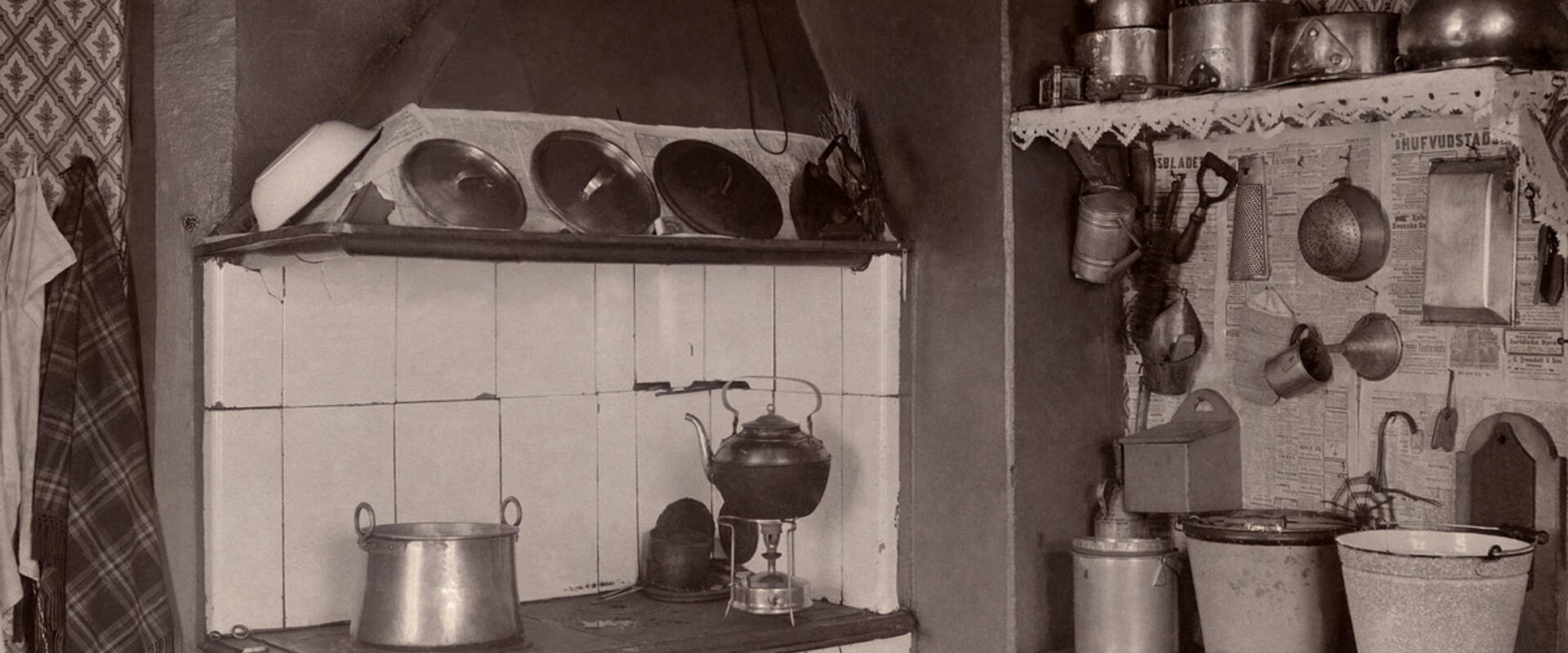 Vasemmalla on puuhella, oikealla on senkki, jonka päällä on sankoja. Seinällä on keittiövälineitä ja seinällä kattilahylly.