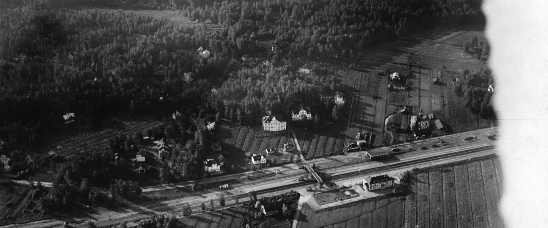 Keskellä kuvaa kulkee junarata, sen ympärillä on rakennuksia mutta enimmäkseen metsää ja peltoja. 