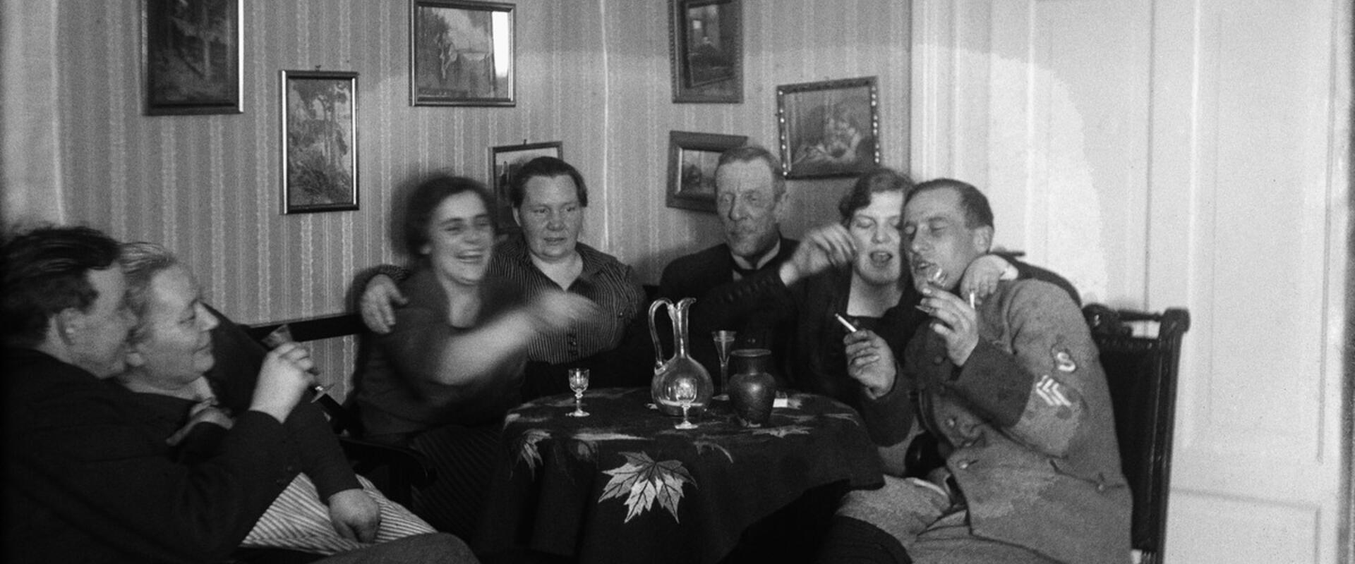 Seitsemän ihmistä istuu pyöreän pöydän ääressä. Joillakin on kädessä savuke ja snapsilasi. Pöydällä on snapsilaseja ja kaksi juomakaadinta. Yhdellä miehistä on suojeluskunnan univormu.