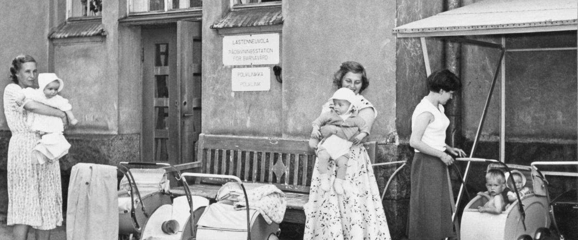Kesämekkoihin pukeutuneita äitejä, pieniä lapsia ja lastenvaunuja Auroran sairaalan pihalla.